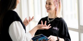 Das Foto zeigt zwei junge Frauen: eine Lehrerin und eine Auszubildende die miteinander interagieren. Die Auszubildende hält ein Pflege-Buch in der Hand.
