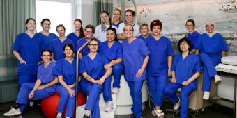 Gruppenfoto des Hebammen- und Pflegeteams des Kreißsaals der Asklepios Klinik Nord in blauer medizinischer Arbeitskleidung innerhalb eines Behandlungs- und Aufenthaltsraums.